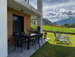 An outdoor patio with a table and chairs, a potted plant, and a view of a green field and mountains. - Apartment 'Tommaso' in Villa Flaminia, Colico, with Garden and BBQ (Colico)