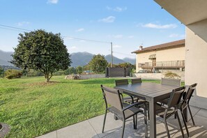 An outdoor patio with a table and chairs, surrounded by a grassy lawn and a tree, with mountains in the background.