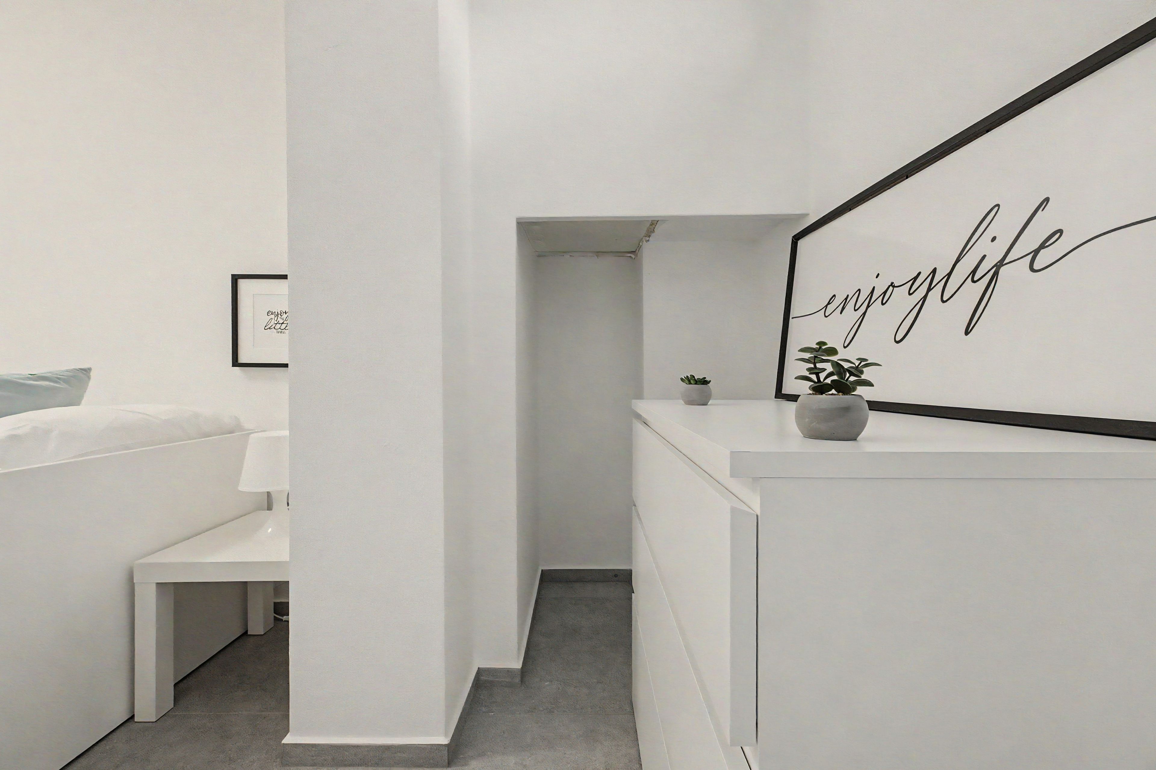 Un dormitorio moderno y minimalista con cama, mesita de noche y un espejo montado en la pared con la frase 'enjoy life' en cursiva.