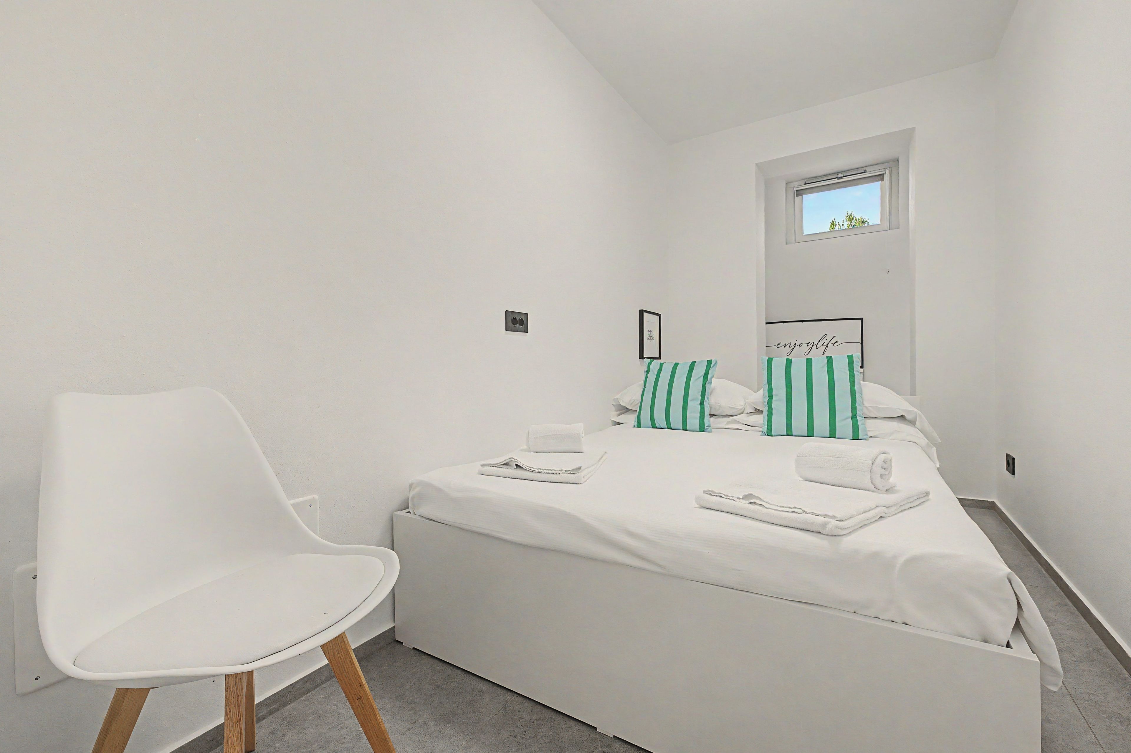 Una camera da letto piccola e bianca con un letto singolo, una sedia bianca e una finestra che si affaccia sul cielo.