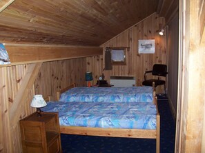Une chambre avec un plafond en bois, un lit recouvert d’une couvre-lit à motifs bleus, une table de chevet avec une lampe, une chaise et une petite table avec une horloge.