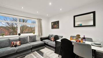 A modern living room with a grey sofa, a dining table with chairs, a large window with a view of buildings, and a painting on the wall.