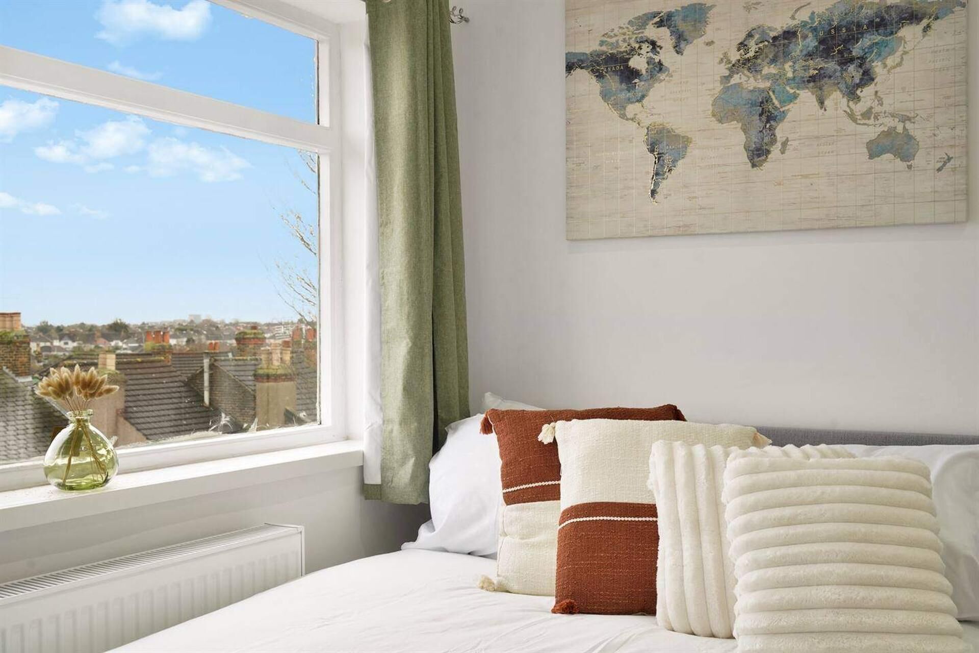 Een slaapkamer met een bed, kussens, een wereldkaart aan de muur, een raam met uitzicht op gebouwen, en een radiator.