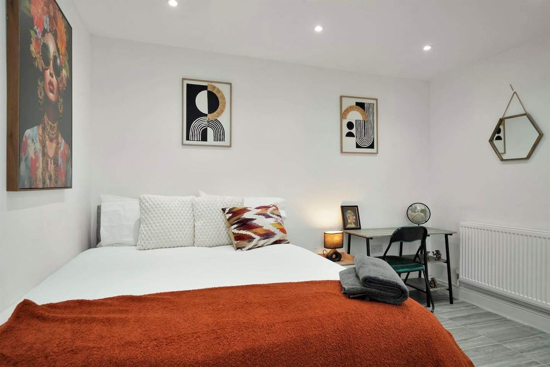 A bedroom with a bed, a desk, and a chair. There are wall art pieces and a mirror.