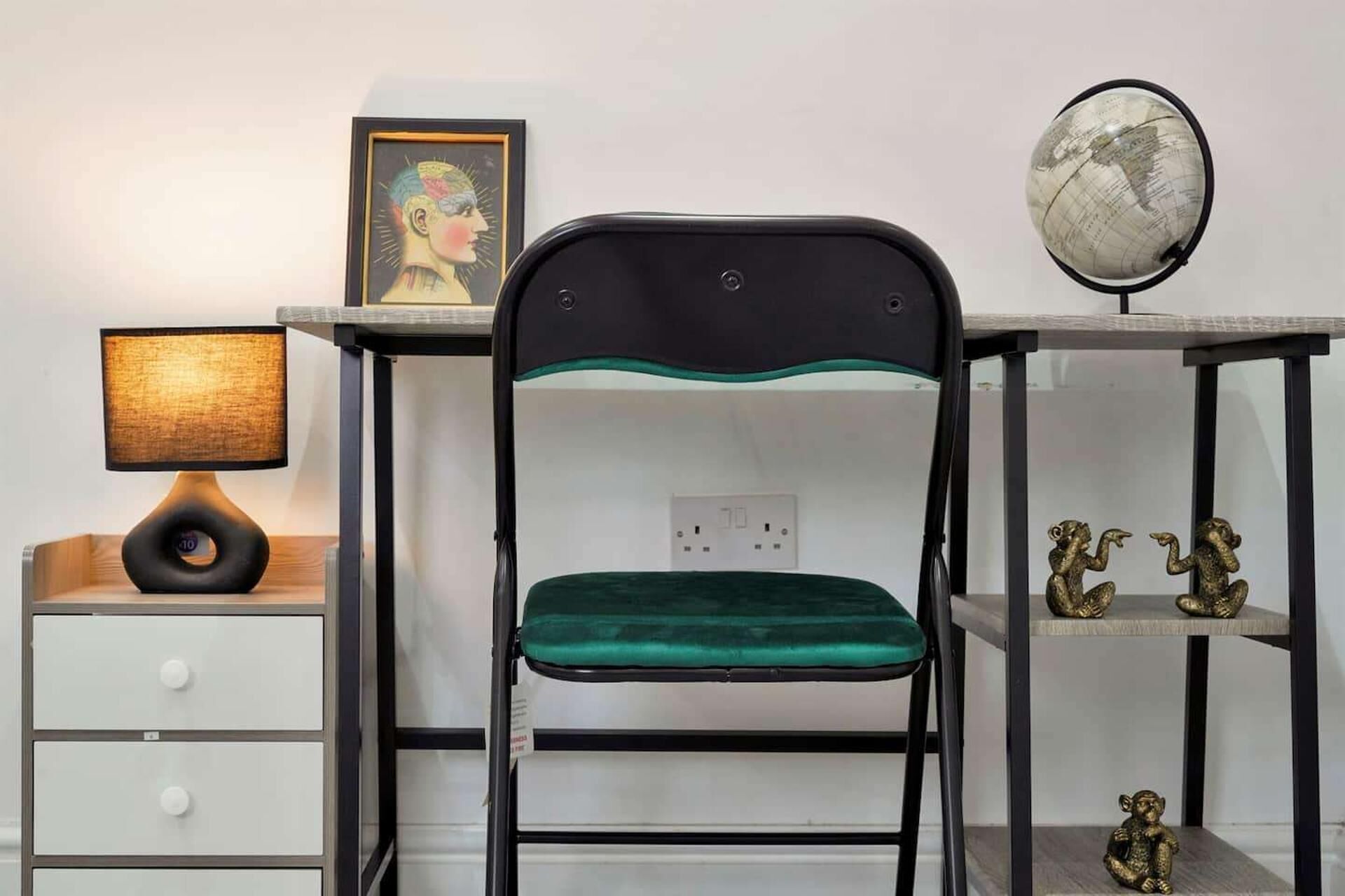 A chair with a green cushion, a desk with a globe and figurines, and a lamp on a bedside table.