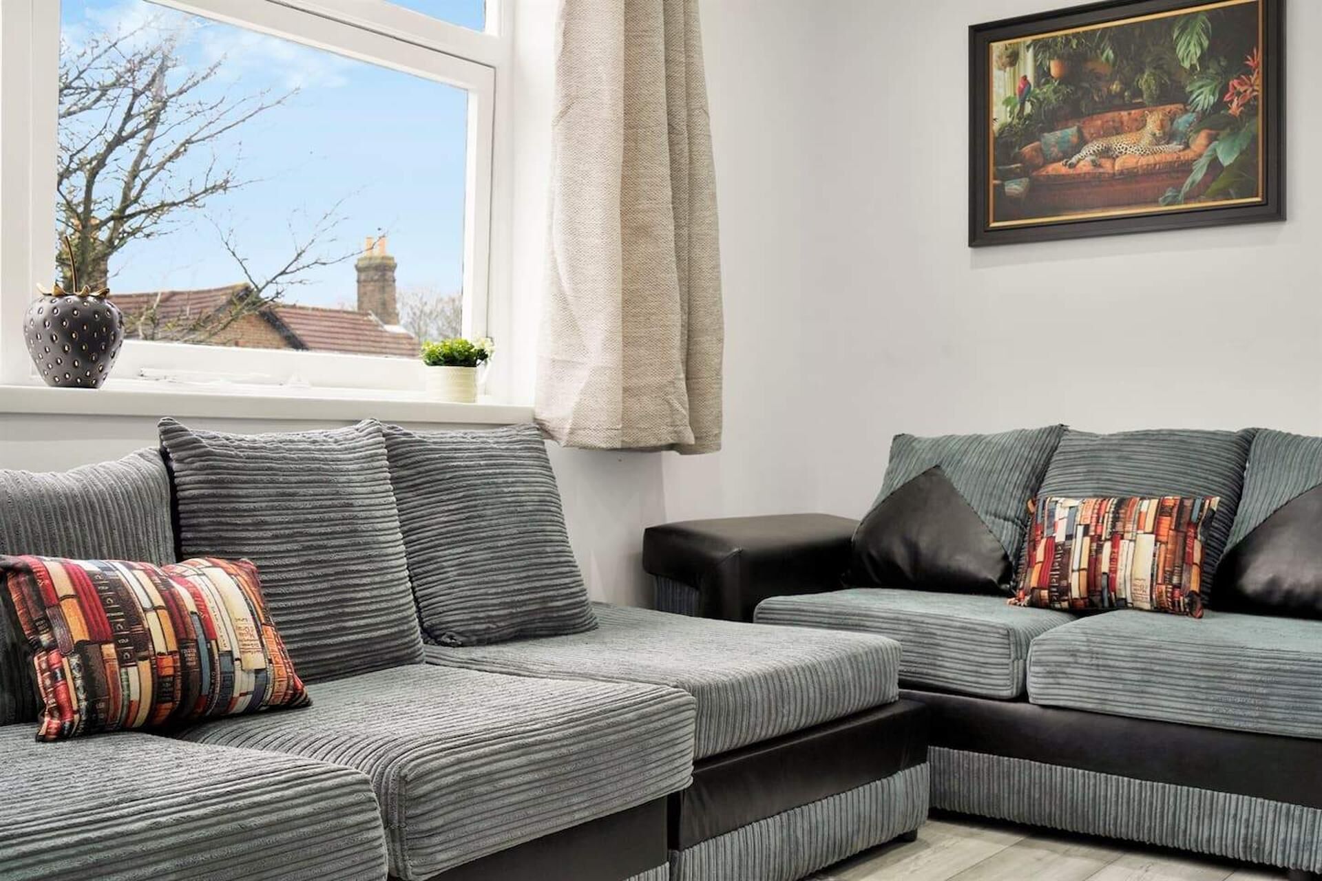 Un salón con un sofá gris, un reposapiés negro y una ventana que da a vistas de árboles y edificios.