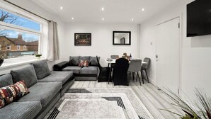 A modern living room with a sofa, a dining table with chairs, a flat-screen TV mounted on the wall, and a window with a view of a house. - Belvedere 4-bed Apt, Sleeps 9 With Parking (Belvedere)