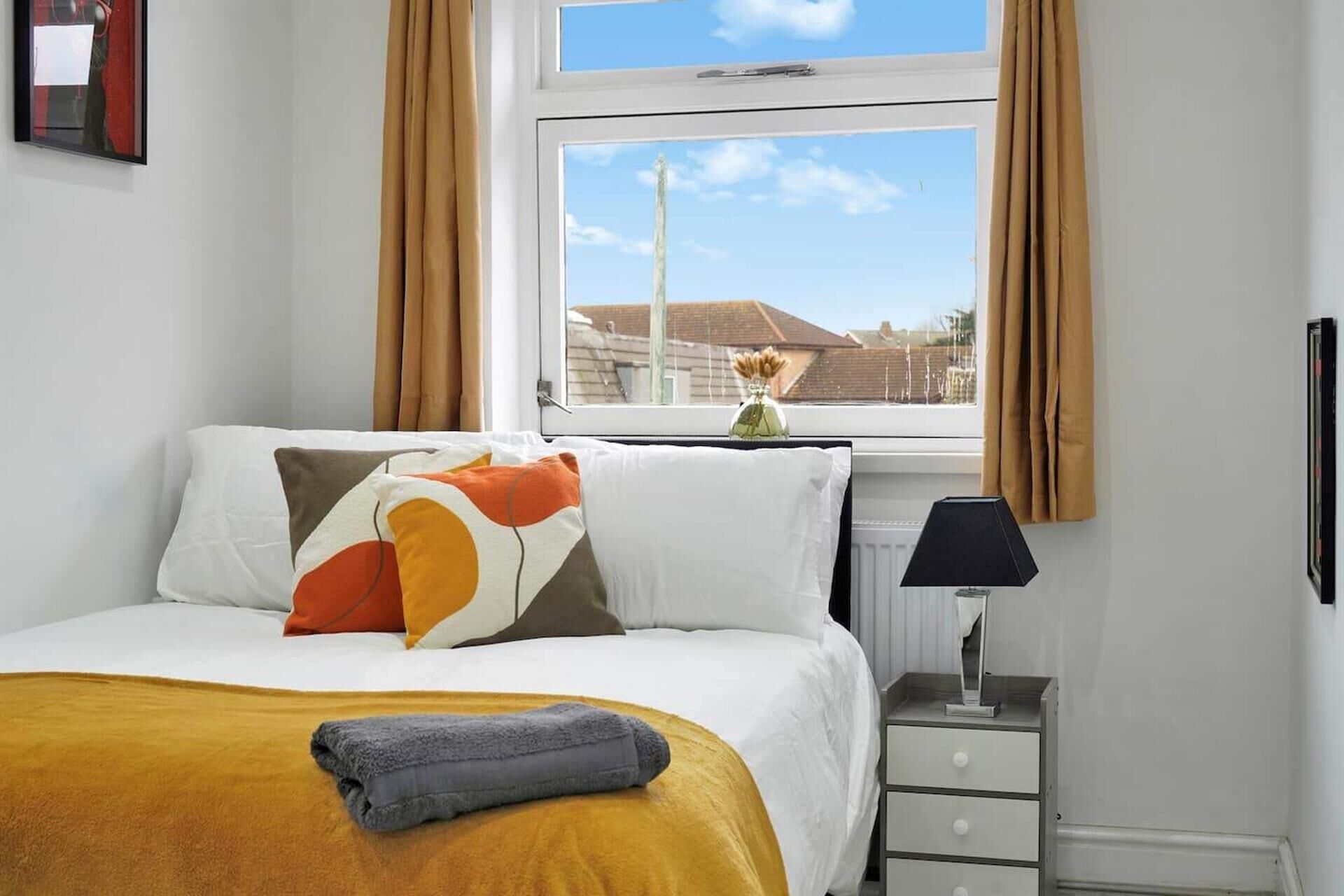 A hotel room with a bed, a bedside table with a lamp, a window with curtains, and a view of the outside.