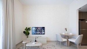 A modern hotel room with a flat-screen TV, a small dining table, two chairs, a white sofa, a plant, and a kitchen area. - Bloomfields Studio in Fairways 3  (Abu Dhabi)
