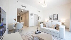 A modern living room with a white sofa, a dining area with chairs, a TV, and a chandelier. - Bloomfields Studio in Fairways 3  (Abu Dhabi)