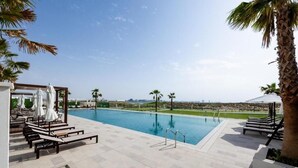 A swimming pool with lounge chairs and umbrellas, surrounded by palm trees and a clear sky. - Bloomfields Studio in Fairways 3  (Abu Dhabi)