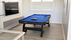 Une table de billard recouverte d’un drap bleu, un téléviseur fixé au mur et une fenêtre avec des stores.