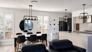 Une cuisine moderne dotée d’une cuisinière intégrée, d’un coin repas avec une table ronde et des chaises, et d’une grande fenêtre avec des stores.