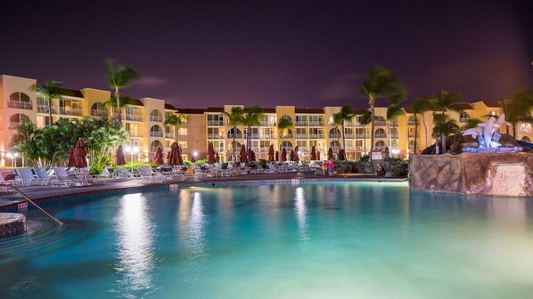 Uma área de piscina de hotel com espreguiçadeiras e guarda-sóis, uma piscina grande e um edifício com várias varandas.