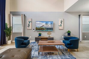 Ein modernes Wohnzimmer mit einem Flachbildfernseher, zwei blauen Sesseln, einem Holztisch und einem gemusterten Teppich.