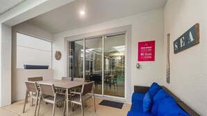 Un patio couvert avec une table et des chaises, une porte vitrée donnant accès à une piscine, et un panneau indiquant les règles à respecter au bord de la piscine.