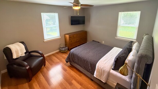Quarto com cama, uma cadeira, cômoda, ventilador de teto e duas janelas com persianas.