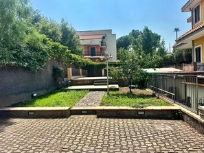 A paved courtyard with a metal fence, a small tree, and a building with a red roof in the background. - Villa dell'Etna - Country Residence - (Mascalucia)