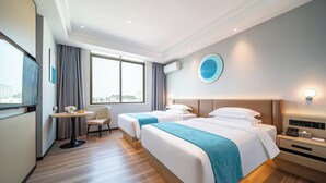Quarto de hotel com duas camas, uma escrivaninha, uma cadeira, um espelho redondo e uma janela com vista para a cidade.