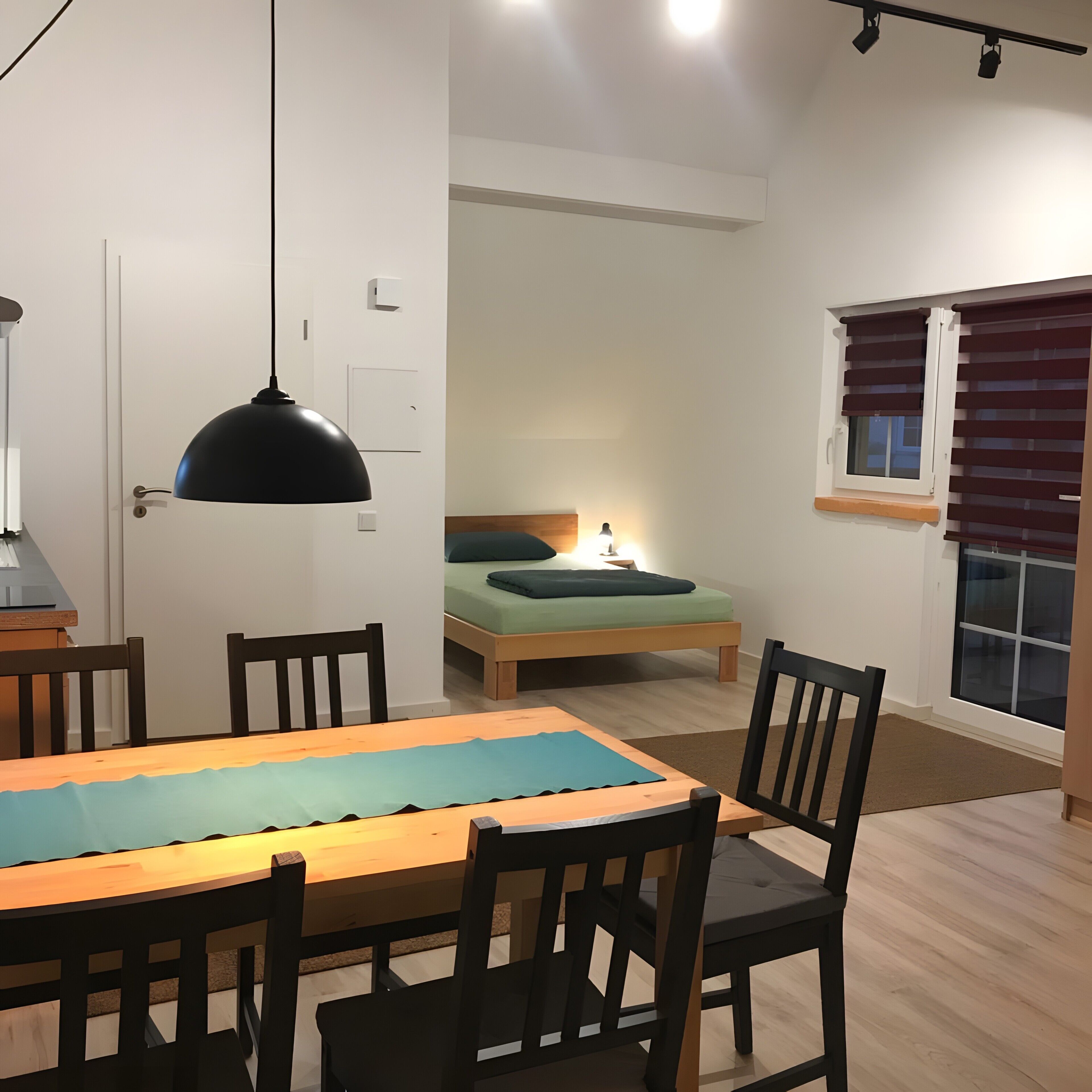 A compact living space with a dining table, chairs, a bed, a window with blinds, and a ceiling light fixture.