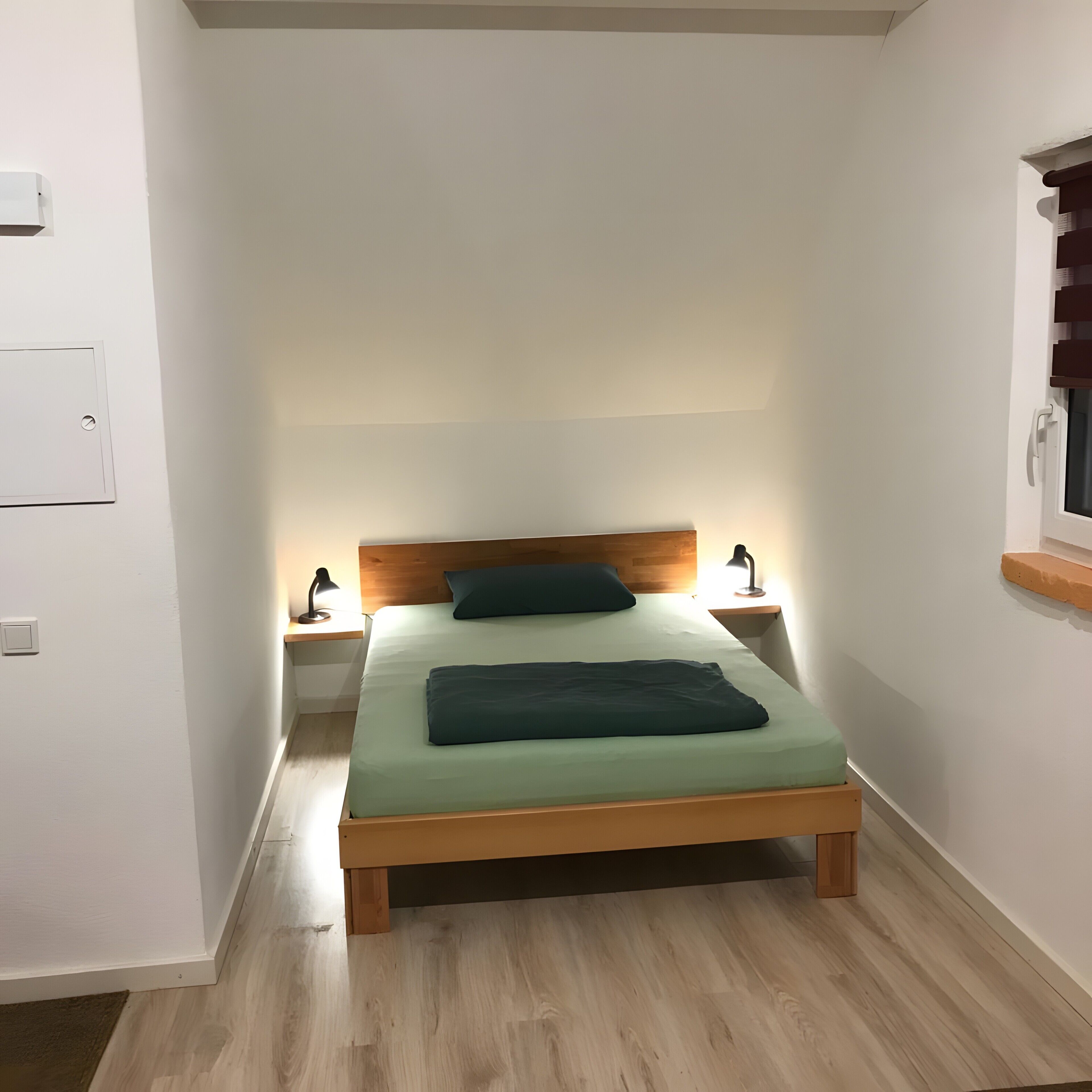 Habitació petita i estreta amb un llit individual, dues taules de nit amb làmpades i un panell de fusta al capçal.