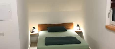 Une petite pièce étroite, avec un lit simple, deux tables de chevet équipées de lampes et une tête de lit en bois.