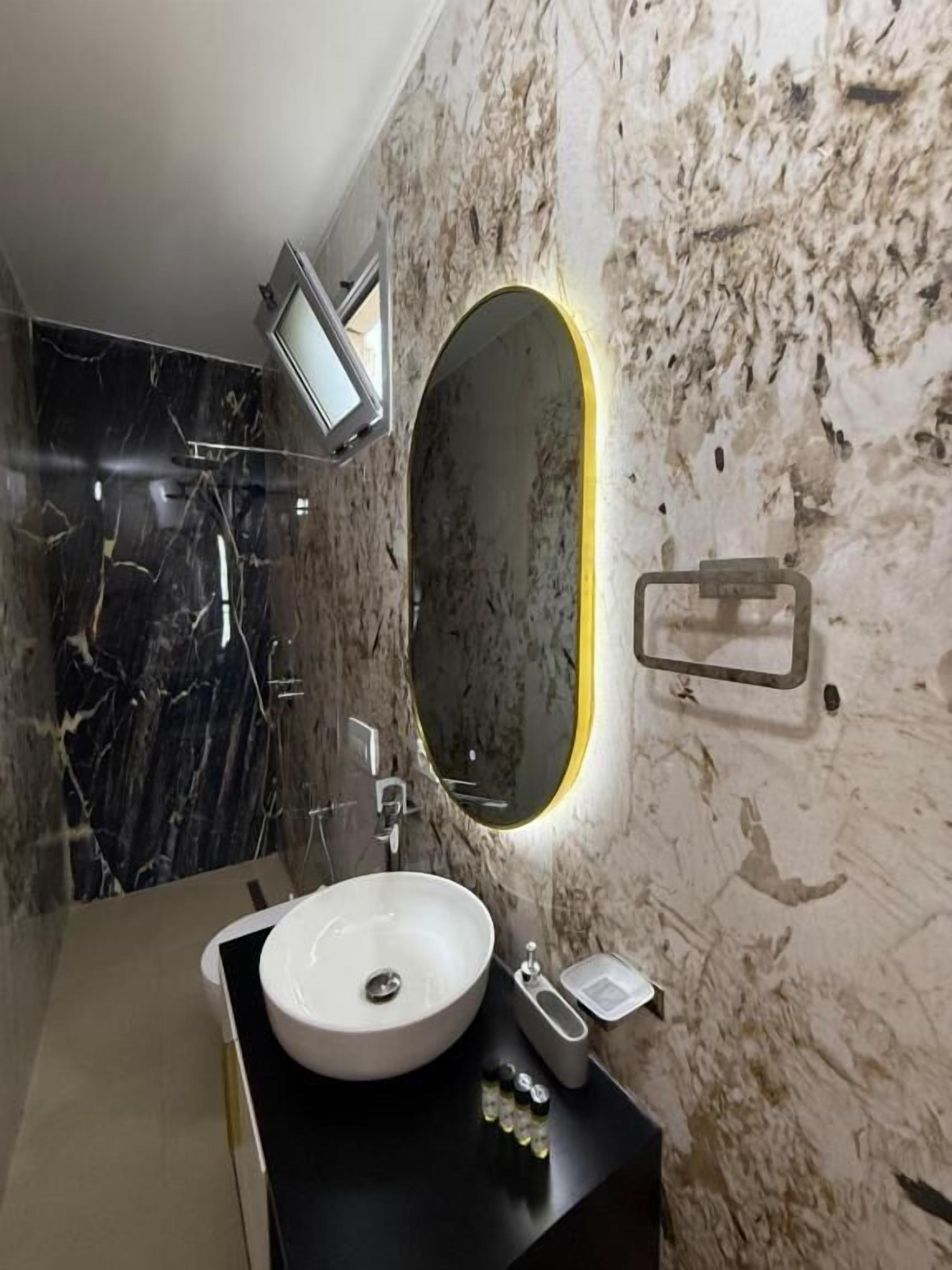 Une salle de bain avec un lavabo en marbre, un miroir éclairé d’une lumière jaune et un miroir à main.