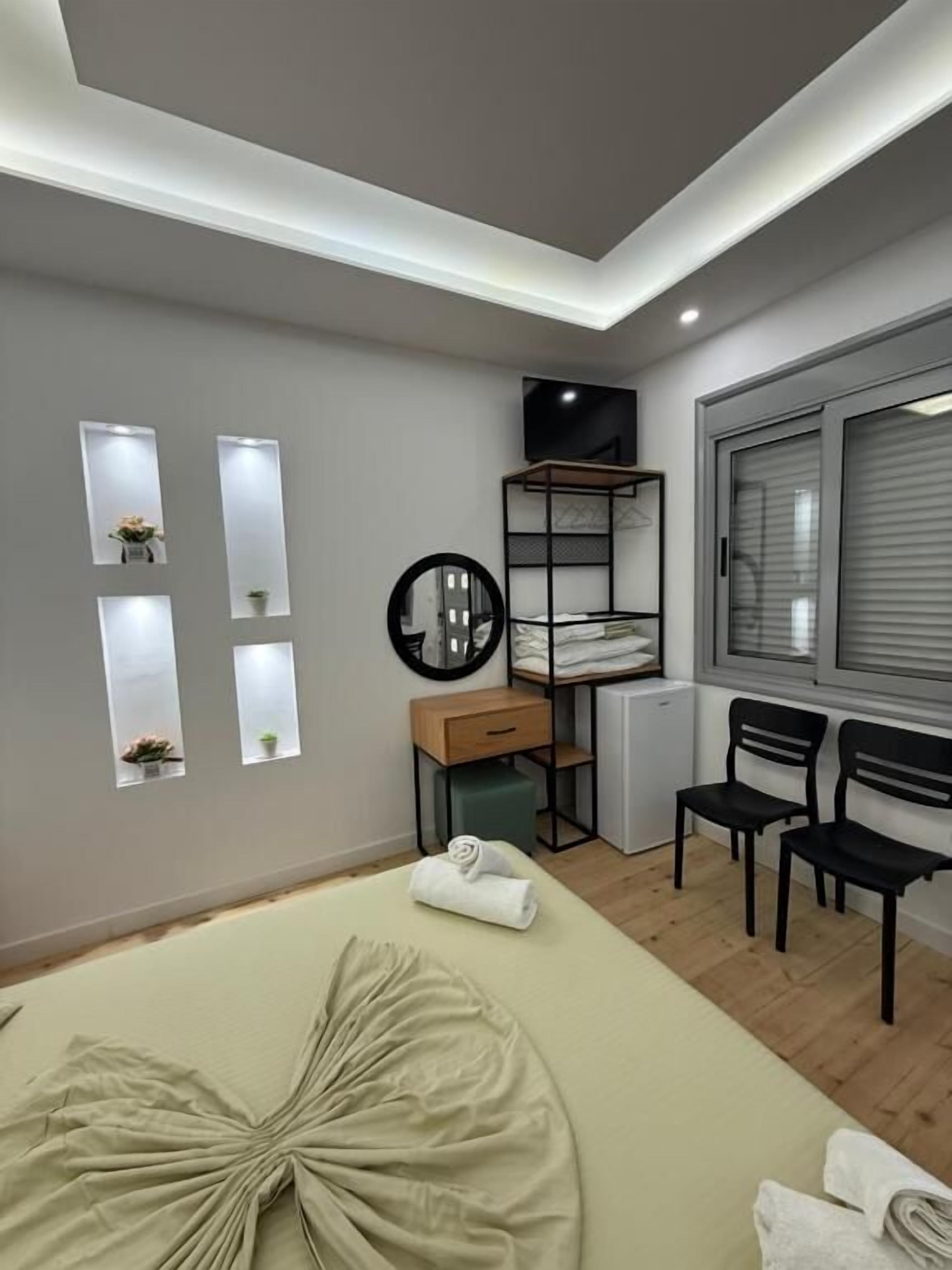 A modern bedroom with a bed, a desk, two chairs, a radiator, and a window with blinds.