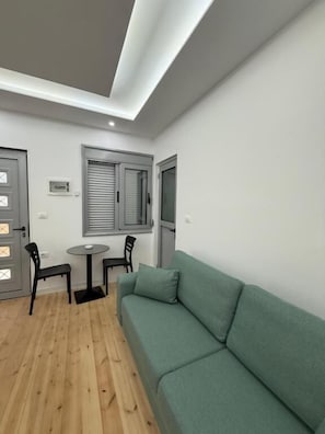 A modern living room with a green sofa, a small round table, two chairs, and a window with blinds. - Jofri Rooms (Himarë)
