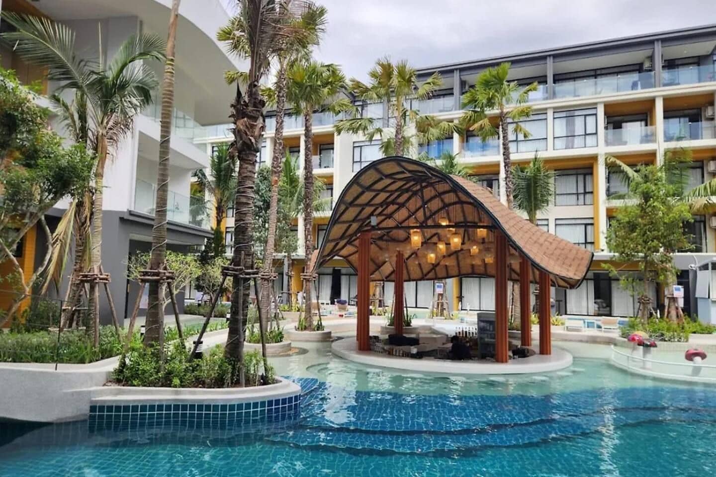 A hotel pool area with a covered seating area, palm trees, and a multi-story building in the background.