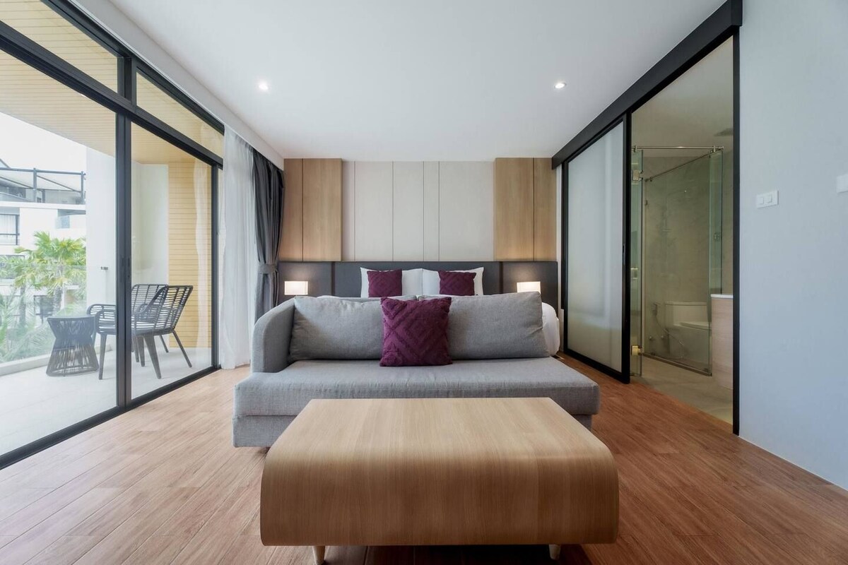 Quarto de hotel moderno com um sofá cinzento, um banquinho de madeira e box de chuveiro de vidro.
