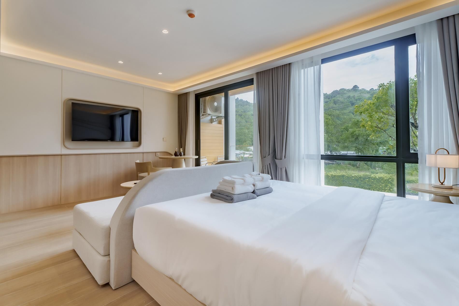 Habitación de hotel moderna con una cama grande, televisor de pantalla plana montado en la pared, una mesita con sillas y un ventanal con vistas a zonas verdes.