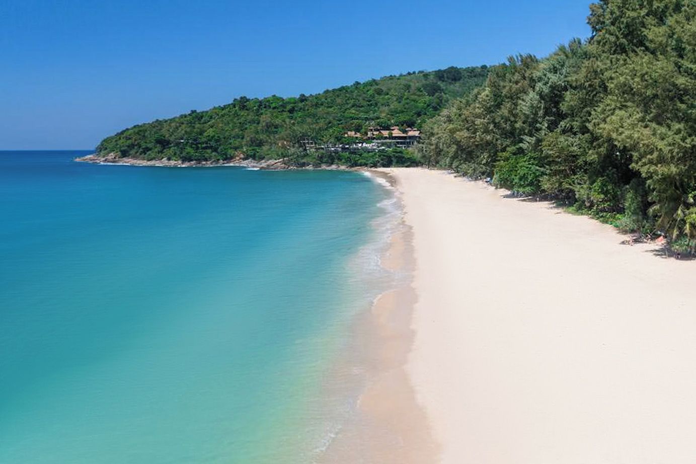 Una playa con agua turquesa cristalina, orilla arenosa y una colina boscosa al fondo.