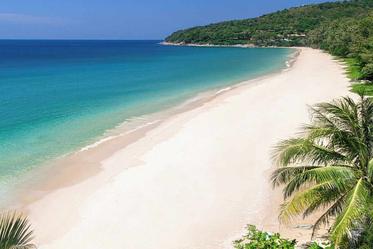 Uma praia com águas turquesa cristalinas, areia branca e uma colina arborizada ao fundo.