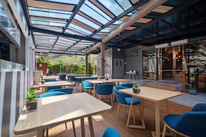 Un espace de restauration intérieur et extérieur avec des tables en bois et des chaises bleues, un plafond en verre et une vue sur la verdure.