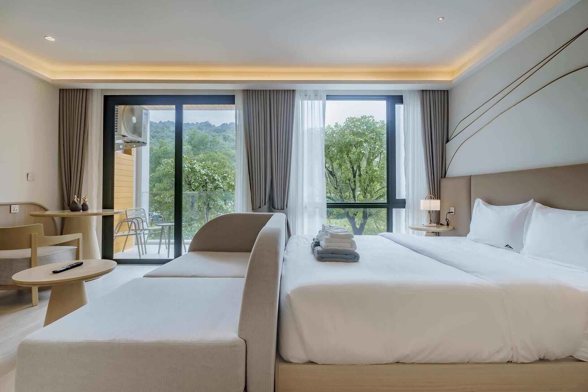 A hotel room with a large bed, a smaller bed, a sofa, a table, and a chair. There is a balcony with a view of trees and a building.