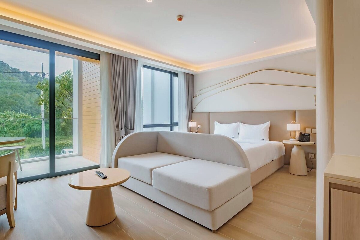 Quarto de hotel com cama, sofá, mesa pequena e vista para o verde lá fora.