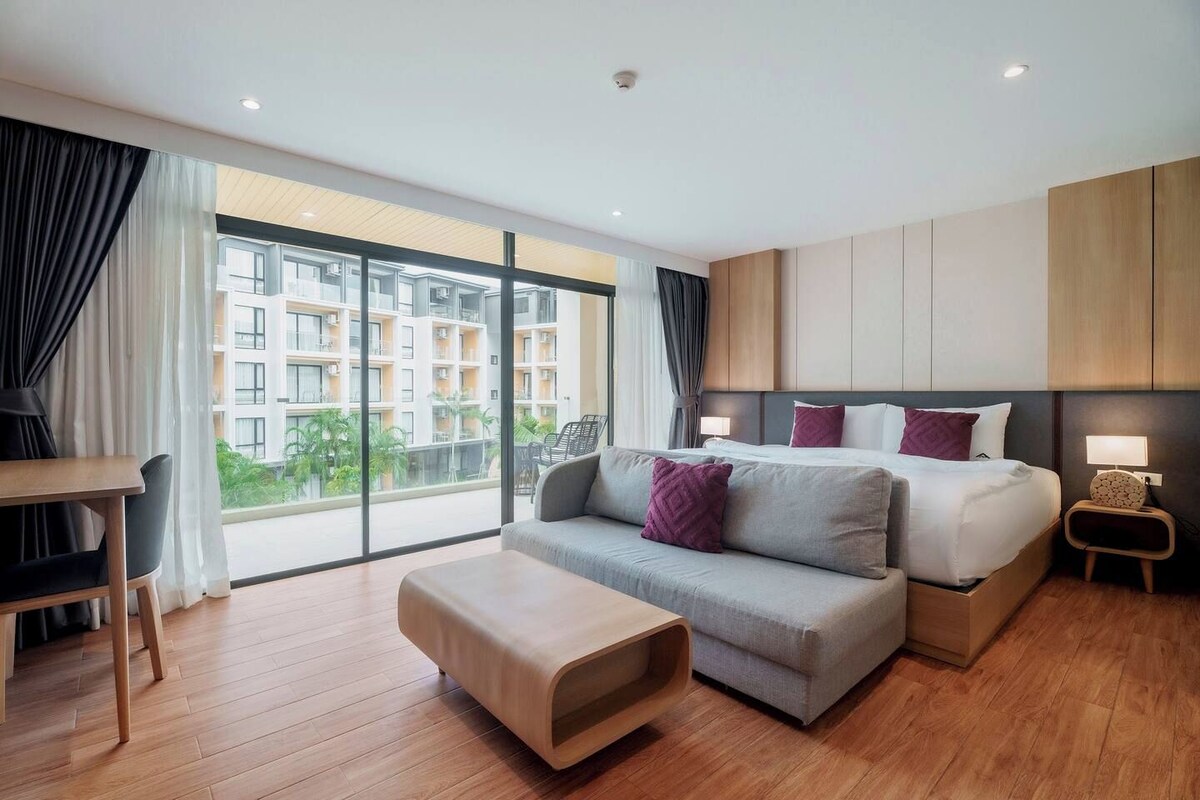 Quarto de hotel moderno com uma cama grande, um sofá, uma mesinha e vista para um prédio residencial.