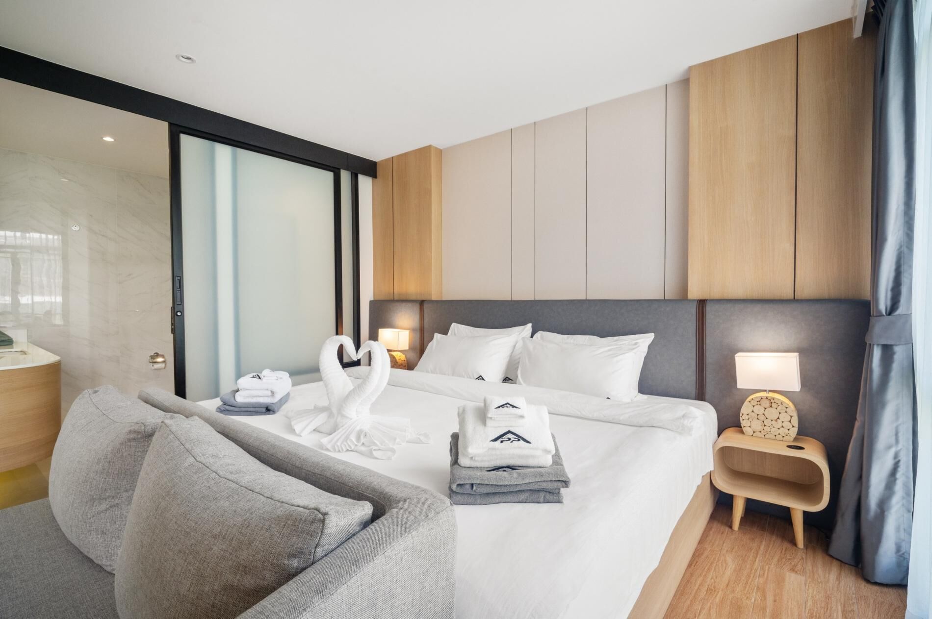 A modern hotel room with a large bed, a sofa, a nightstand, and a glass door leading to a balcony.
