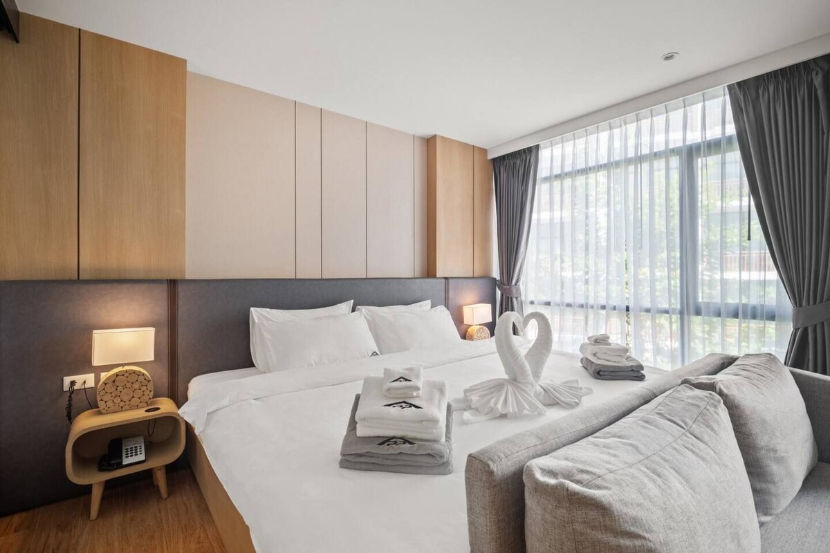 Quarto de hotel moderno com uma cama grande, mesa de cabeceira com abajur, sofá e vista para o exterior através de amplas janelas.