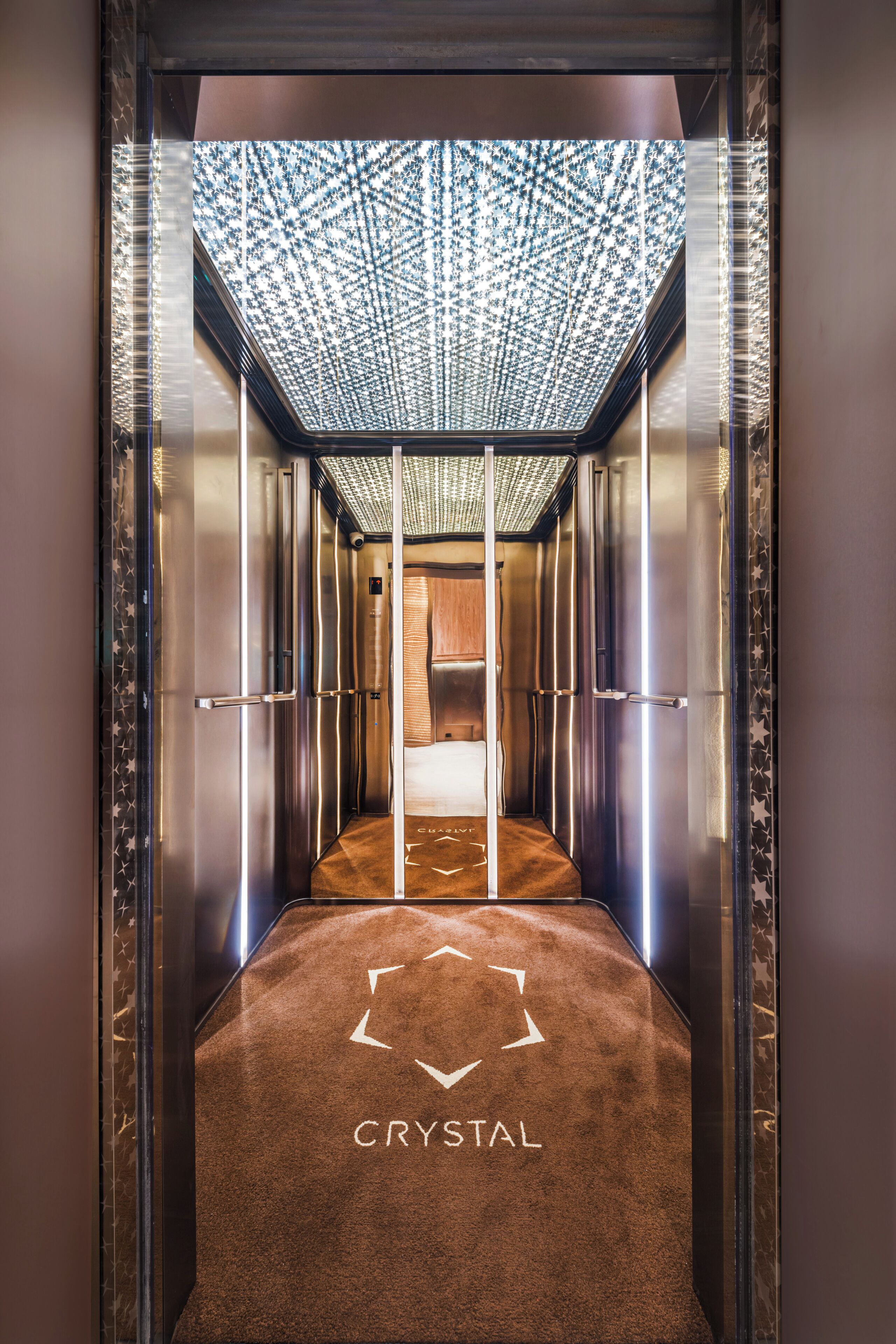 Interior de un ascensor con techo estampado, alfombra con flechas direccionales y la palabra 'CRYSTAL' en el suelo.