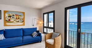 Une pièce avec un canapé bleu, un fauteuil en osier, un tableau accroché au mur et une vue sur la mer, visible à travers de grandes fenêtres.