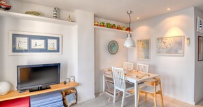 A modern living room with a flat-screen TV, white dining table, and chairs, a bookshelf with decorative items, and a ceiling light. - Seafront apartment (Calella de Palafrugell)