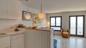 Een moderne keuken met een vaatwasser, een eethoek met een blauwe stoel, en uitzicht op zee door grote ramen.