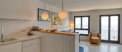 A modern kitchen with a dishwasher, a dining area with a blue chair, and a view of the sea through large windows.