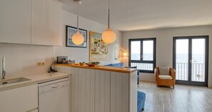A modern kitchen with a dishwasher, a dining area with a blue chair, and a view of the sea through large windows. - Seafront apartment (Calella de Palafrugell)