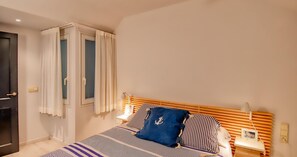 A bedroom with a bed, a nightstand, a lamp, a window with curtains, and a door. - Seafront apartment (Calella de Palafrugell)