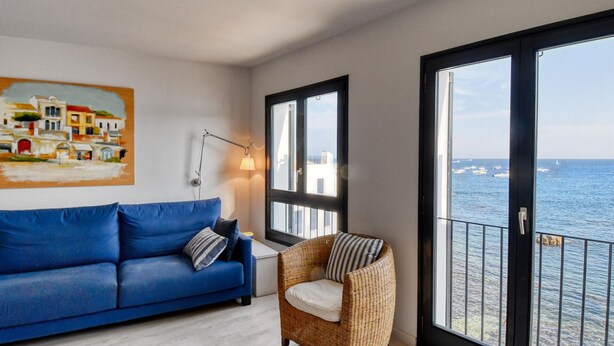 Een kamer met een blauwe bank, een rieten stoel, een schilderij en uitzicht op de zee door grote ramen.