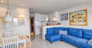 A modern living room with a blue sofa, white dining table, and chairs. A kitchen is visible in the background with white cabinets and a refrigerator. - Seafront apartment (Calella de Palafrugell)