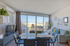 Un salon moderne doté d’une grande fenêtre, d’un téléviseur à écran plat, d’une table à manger avec des chaises et d’un canapé avec des coussins.